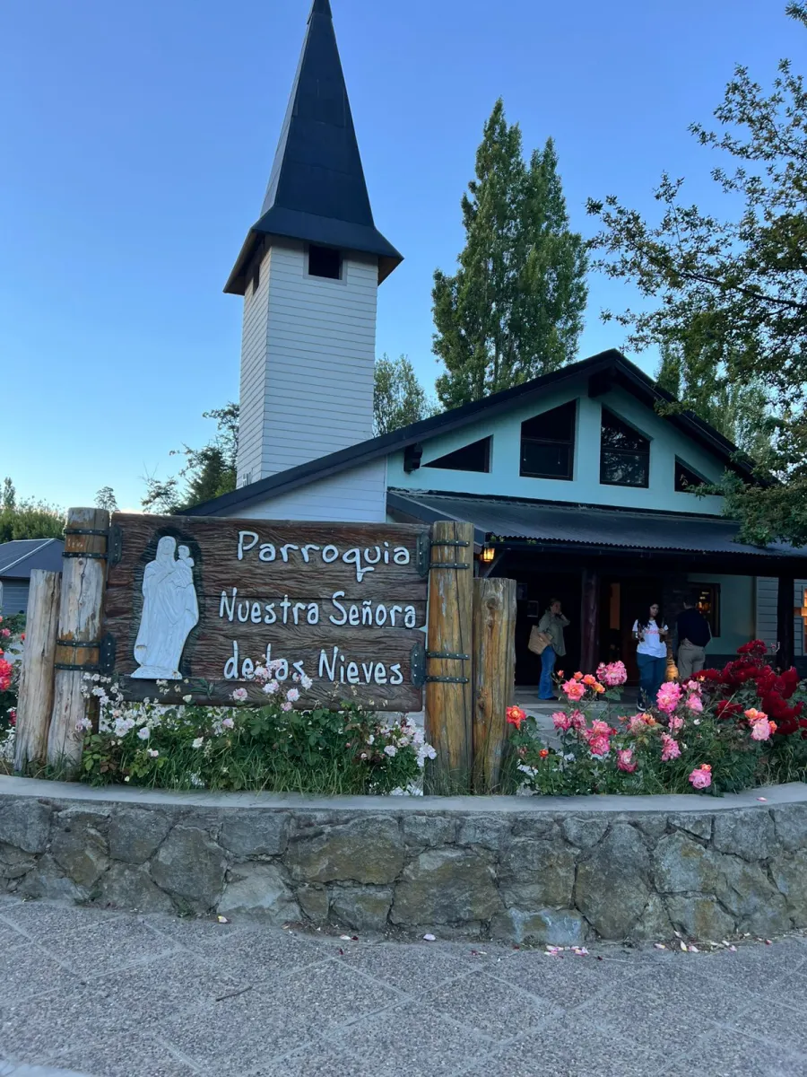 Frente de la Parroquia Nuestra Señora de las Nieves: campanario blanco, cartel de madera y rosales
