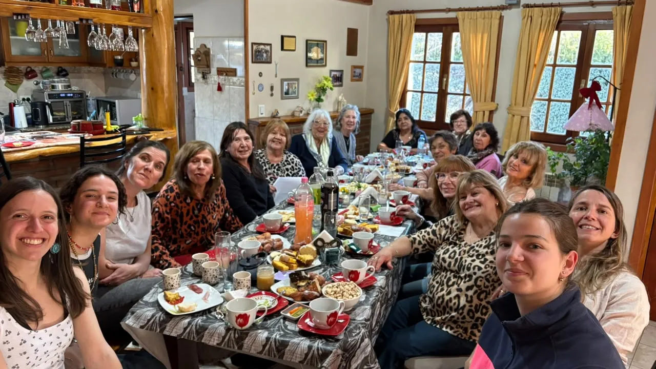 Mujeres de distintas generaciones sonríen alrededor de una mesa larga con tazas, facturas y mate, en una casa con ventanas al jardín